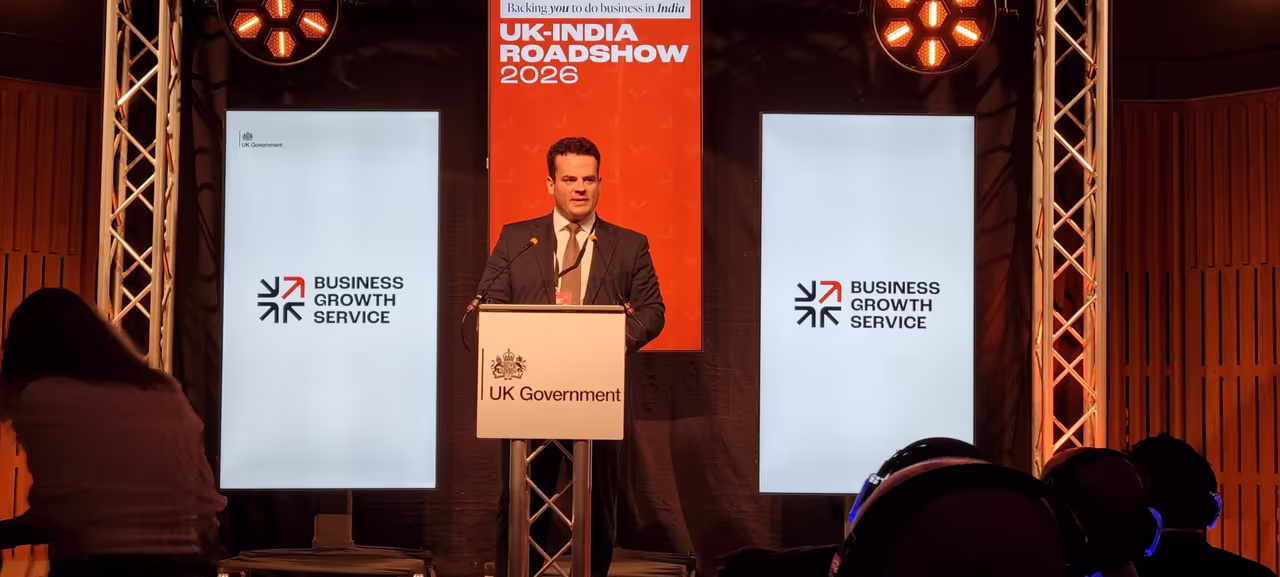 A speaker addressing delegates from the UK Government lectern at the UK–India Roadshow 2026 in Leeds, flanked by Business Growth Service screens and the orange UK–India Roadshow banner